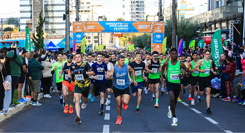 Largada de uma corrida de rua com muitos participantes, passando por um pórtico de largada onde se lê 'Circuito dos Vales'.
